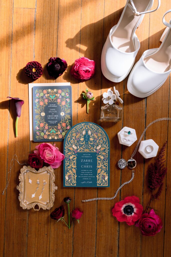 Jewel-toned wedding flatlay featuring invitations, flowers, jewelry, perfume, and bridal shoes arranged on a wooden floor.
