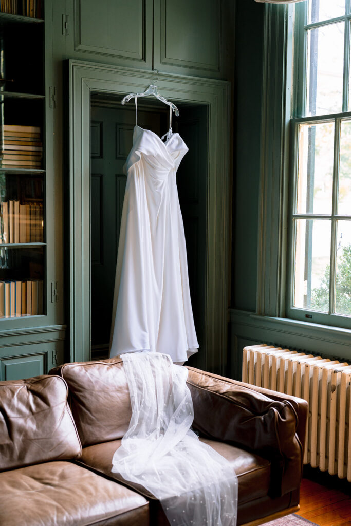Wedding dress hanging on a door inside a historic room with sage green walls, next to a window and a leather sofa with the veil draped over it.