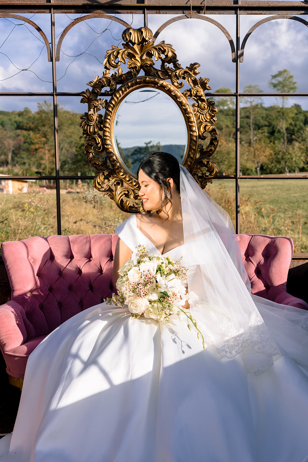 Bride sitting on a pink vintage settee, holding a white bouquet, with a gold ornate mirror behind her and natural light highlighting her dress.