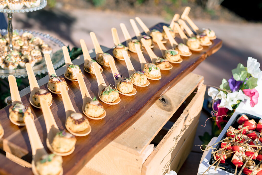 Elegant bite-sized wedding appetizers served on wooden spoons and displayed on a rustic wooden board outdoors.