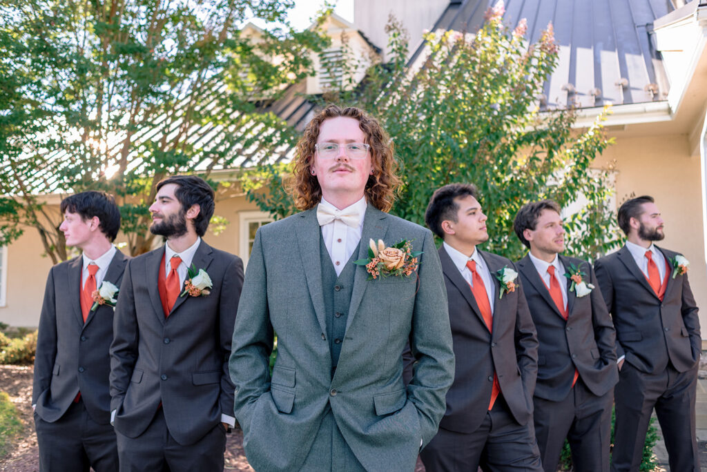 Groom in a green suit standing confidently with his groomsmen behind him, all wearing coordinating suits with orange ties and bold color boutonnieres.