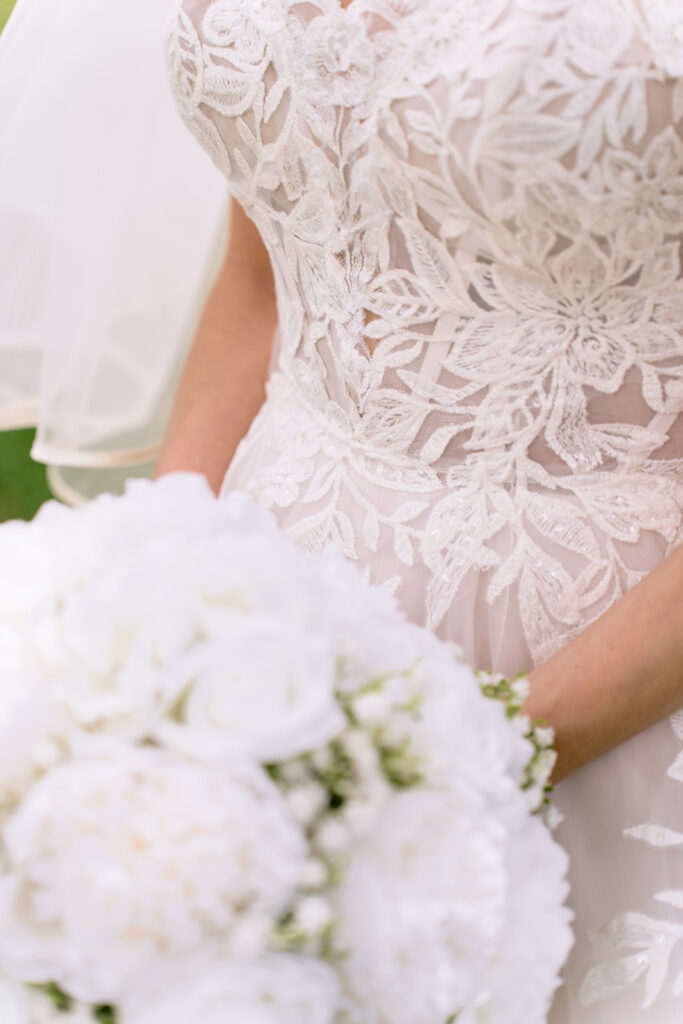 Close up of a bride holding a white peony bouquet, showcasing the intricate lace detailing on her wedding dress.