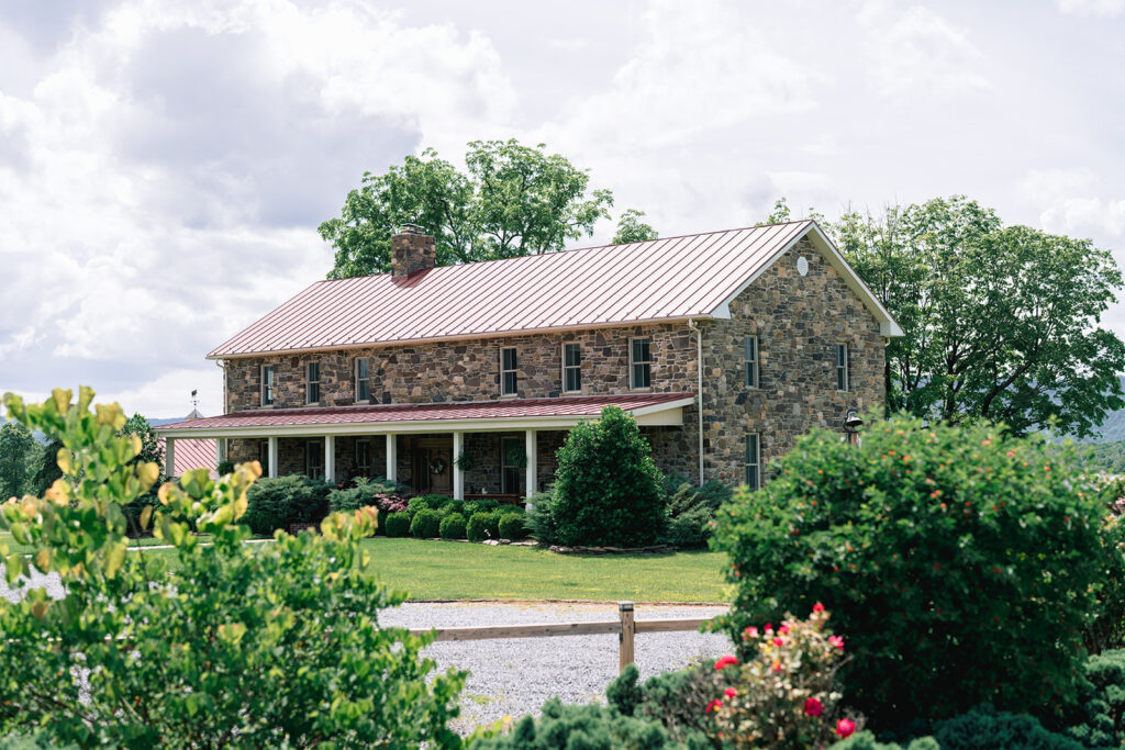 Historic stone farmhouse with a red metal roof surrounded by lush greenery and garden landscaping on a sunny wedding day.
