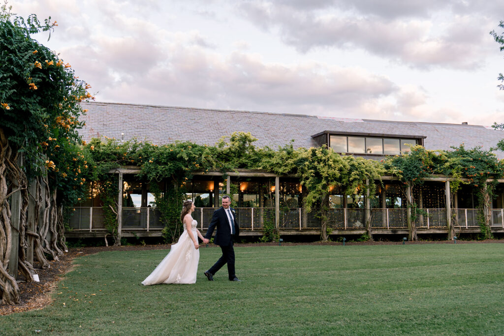 Bride and groom holding hands while walking across a lawn at sunset, with a vine-covered garden venue behind them.