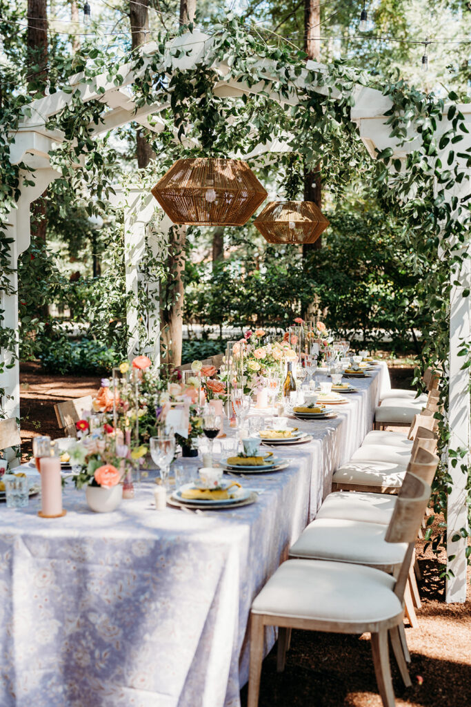 Outdoor wedding dinner under a pergola with greenery, soft florals, and neutral table settings in natural light