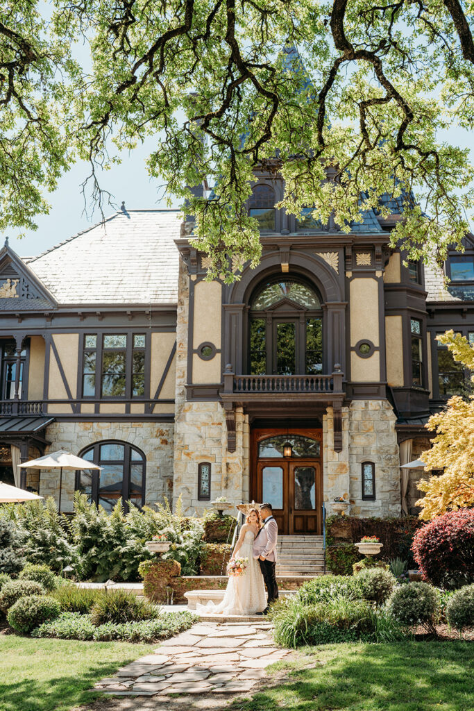 Wedding couple standing together in front of a historic stone estate surrounded by greenery and natural light